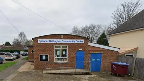 Google Single-storey brick building with "Spencer Dallington Community Centre" on a sign above the main window.  It has blue doors and notices in the window, with a ramp up to the main door. There is a skip to the left and cars parked in the road to the right.