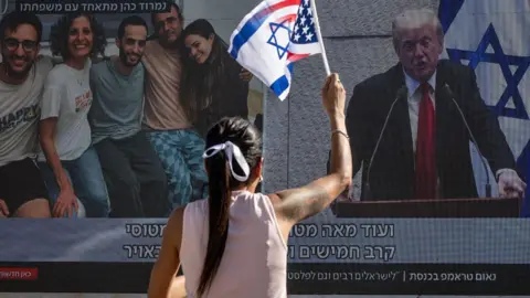 Getty Images A woman waves Israeli and US flags while watching a screen that shows side-by-side footage of released Israeli hostages and US President Donald Trump