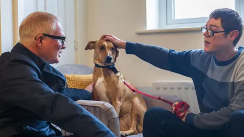 West Midlands Combined Authority Richard Parker on the left wearing black glasses looks at David Hopper who is also wearing black glasses, a blue jumper while patting a gold-coloured dog on the head that is sitting in the middle on a grey chair.