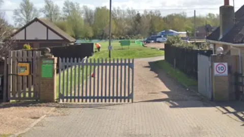 Google Two silver gates are in between two houses. The one on the right is open and a driveway leads to a building. There is a 10mph sign on a pillar on the right and a defibrillator is on the left gate.