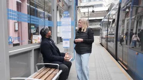 A black man sat a tram stop in a black coat and hat. He is talking to a female BBC reporter with blonde hair. She is wearing a black gillet and faded blue jeans. A blue Midlands Metro tram is passing by