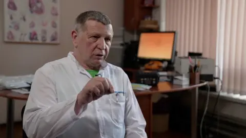 Kevin McGregor / BBC Serhiy Maksymenko sits in his consulation room. He wears a white doctors coat. He was part of the team of surgeons who removed the shrapnel from Serhiy’s heart.