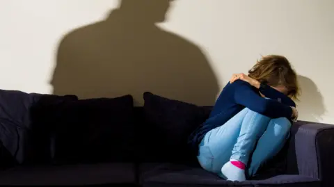 PA Images A woman in a blue jumper and jeans cowers, with her face in her hands, in the corner of a navy sofa while a shadow of a man towers above her.