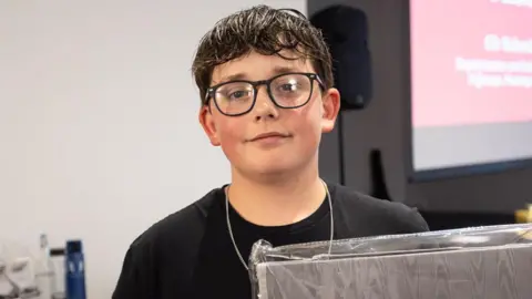 Matthew smiling at the camera, while standing in a room with a screen and a speaker behind him. He has short dark hair and glasses and is wearing a black T-shirt.