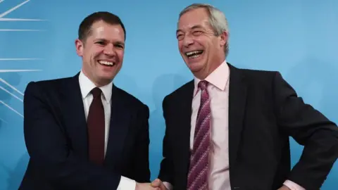 Former British Conservative Party member Robert Jenrick, dressed in a navy suit with a maroon tie, shakes hands with Reform UK leader Nigel Farage, dressed in a black suit with a purple-striped tie and pink shirt, at a press conference. The photo captures the pair mid laugh.