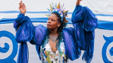 MĀDŁY A woman wearing a sequn top draped in blue fabric and a colourful crown