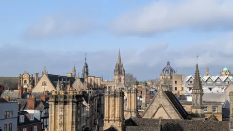 Lucie Johnson The rooftop spires of Oxford in the city centre. The stone buildings are lit up by the sun. The sky over head has some clouds but is mostly blue.