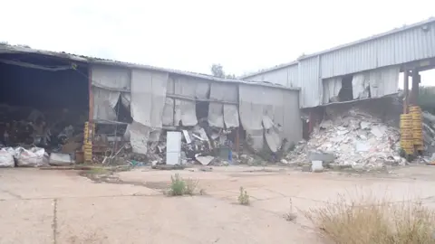 Rubbish and waste can be seen piled up in very dilapidated metal storage buildings with sections missing. The site looks unkempt and abandoned.
