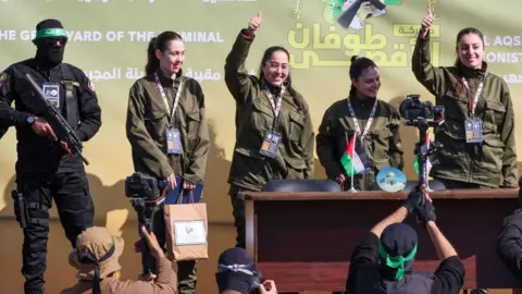 Getty Images A Hamas fighter, armed with a gun, with his face covered, dressed in black, with a green bandana, stands on the left of the stage next to four Israeli female soldiers - two of which hold their hands up with their thumb up - after they were released from being hostages