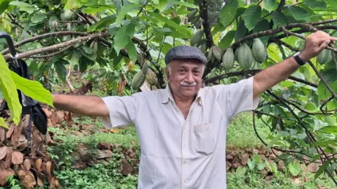 George Matthew George Matthew usando um boné achatado está em frente a um cacaueiro, com frutos de cacau pendurados nos galhos.