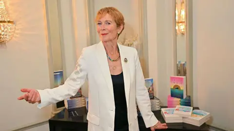 Getty Images Actress Celia Imrie stood in front of a pile of her books. She is wearing a white jacket and black shirt. Her first arm is slightly raised and she is pointing her index finger.