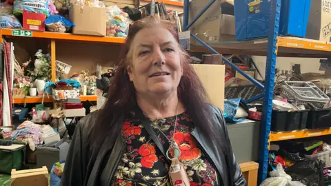 Sameena Akhtar, wearing a black leather jacket and flowery top, stands next to shelves full of goods.