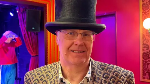 A man wearing a brown and blue blazer with a white shirt that has butterflies all over it and a black top hat.