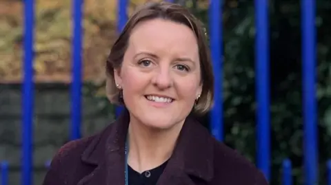 Róisín Blackery is standing beside blue school gates. She has short brown hair and is wearing a purple overcoat and black top. 