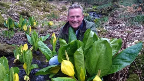 Escape to the Country host Jules Hudson squats behind a plant 