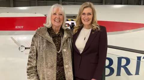 Two women pictured by a Concorde plane at Aerospace Bristol. Marion has short white hair and is wearing a grey coat. Julie is stood with her arm around her. She has long blonde hair and is wearing a white top and burgundy blazer. Both of them are looking at the camera and smiling.