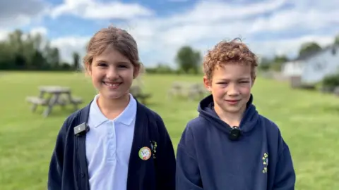 Two children are in a field with picnic benches behind them. On the left the girl has brown hair tied back and is wearing a white polo shirt with a navy cardigan. The boy on the right has short curly dark blond hair and is wearing a navy hoody.