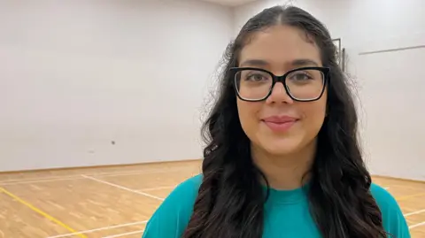 Hannah, a 15-year-old girl, stands in a white-walled school gym and looks into the camera. She has long black hair and is wearing an aquamarine T-shirt and large, black-rimmed glasses.