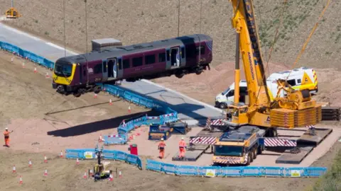 A giant yellow crane in a field next to railway line. It is lifting a train carriage. A temporary track has been built to get the crane into position in the field. There are about a dozen people in orange overalls.
