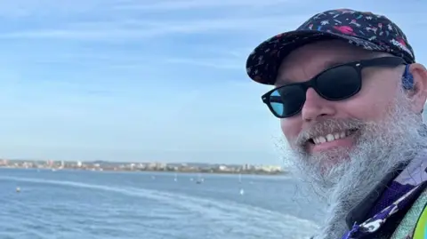 Benjamin Franklin Wells-Lane has a white beard and is wearing a cap and sunglasses.