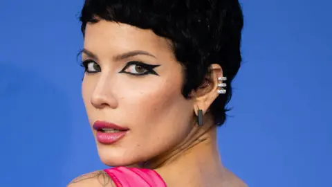 Halsey - a 30-year-old woman with a dark pixie cut. She has brown eyes and wears a graphic black eyeliner, with two wings. She has a sparkly pink lipstick, matching a vibrant pink dress. She looks back at the camera over her left shoulder and is pictured against a blue backdrop..