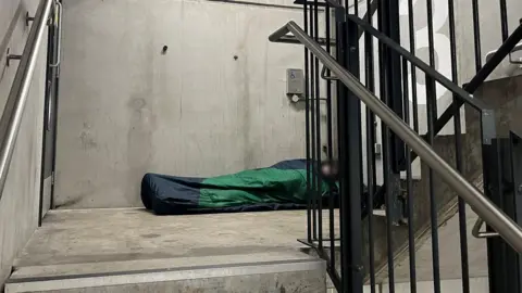 BBC A concrete stairwell inside a car park. A black and silver metal railing obscures the face of a man who is laid on the floor inside a black and green sleeping bag.