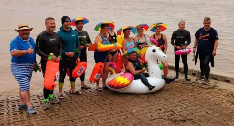 Sarah Scholes Twelve men and women stood in front of the Humber on the pavement wearing swimming uniform. Some of the swimmers have multi-coloured umbrella hats on their head. One swimmer is sat in a unicorn inflatable. 