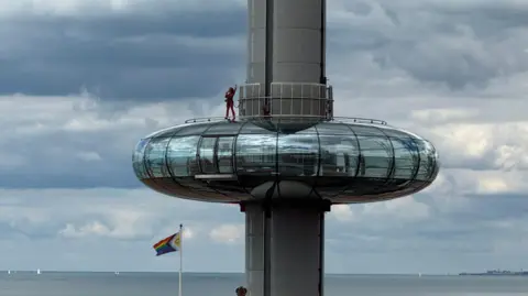 Anthony Martin / BBC A person stood on top of a large circular glass viewing pod. The pod is raised in the air along a large metal pole. The pod is in front of the sea.