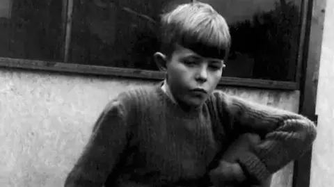 David Bowie Estate A black-and-white photograph of a young David Bowie with short hair, leaning against a wall and looking towards the camera, wearing a long-sleeved jumper.