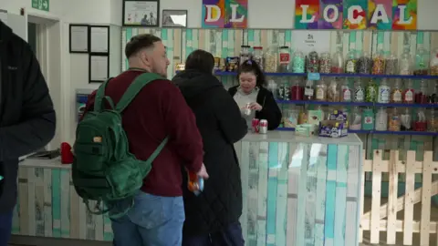 Two people stood in a queue in the shop buying cans of soft drinks. One woman is wearing a dark coat, a man is wearing a purple jumper and a green backpack. A sign about the counter says DL Local.