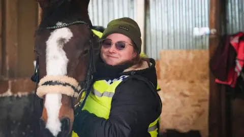 Person in a high-visibility jacket holding a brown horse with a white blaze inside a stable.