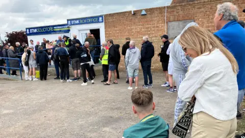 Nadira Tudor/BBC The queue of supporters seen from the car park. It features lots of people winding their way to the ticket office building. In the foreground a woman is looking at a younger man who is sitting on a camping chair.