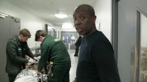 Clive Myrie at a hospital corridor with a bed on the background