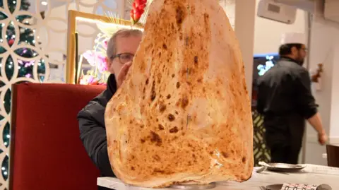 Reporter David Sillito nearly concealed by a giant hanging naan bread at a restaurant