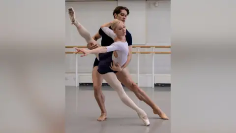 Andre Uspenski Melissa Hamilton dancing with Lukas Braendsrod in a dance rehearsal studio. Melissa is wearing a white leotard, white tights and white ballet shoes. Her hair is blonde and slicked back Lukas is wearing black t-shirt and shorts. He has brown hair. Melissa's right leg is pointed down leaning forward and her left leg is raised behind her. Gareth has his left arm around her waist supporting her and his right arm is supporting her leg.