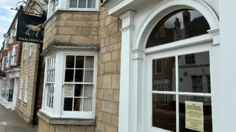 An empty light-stone pub building with white framed windows and a doorway. A sign hanging from the building depicts a white horse and reads Angel and White Horse. In the windows, signs advertising pub management required can be seen.