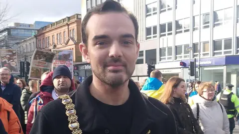 A man dresses in a black coat and a black top is wearing a chain of office and in front of a crowd of people. Some are holding posters and a man in the background is wearing the Ukrainian flag draped over his shoulders. 