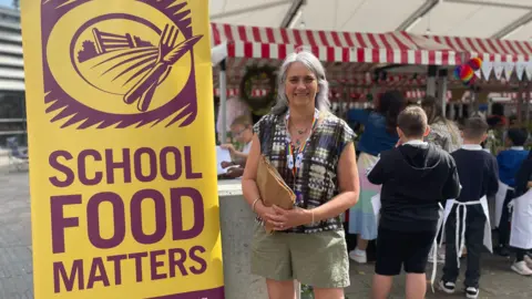 Lisa Didier stood next to a yellow School Food Matters poster. She is holding brown paper bags and is smiling. She is wearing a colourful top and lanyard.
