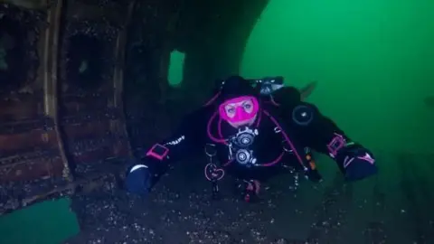 Yvonne Tachley Maddy Kennedy is scuba diving in a dark dry suit and bright pink mask swims inside a sunken metal structure, surrounded by greenish underwater light.