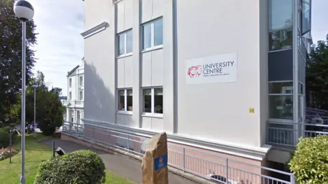 A google street view picture of the outside of the university centre. The building is cream coloured. There are lamp posts outside. 