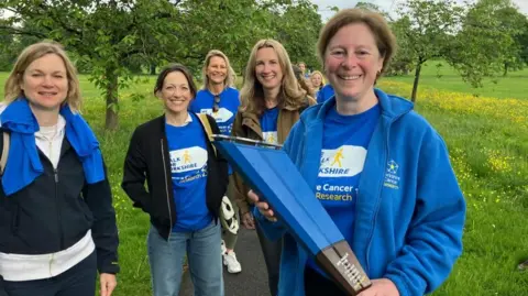 BBC/Seb Cheer A woman, Lisa, holds a large blue baton and smiles at the camera. More than four other women are also in the image, all smiling at the camera. They are in a park, surrounded by grass and trees.