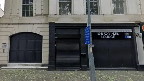 The front of a grey concrete building with large black doors and shutters and a sign reading Decode Lounge above the doors