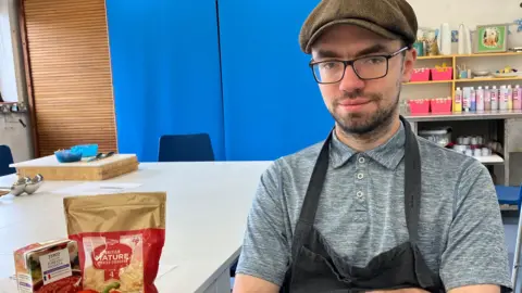 Kate Bradbrook/BBC Man with a flatcap and glasses wearing a grey t-shirt and black apron smiles at the camera. On the table next to him are cooking ingredients including grated cheese and tomato passata