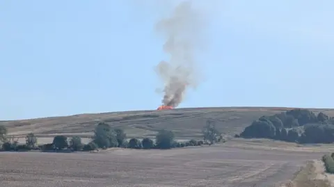 The fire seen from the bottom of the hill at some distance. Orange flames have consumed the hay bales, with lots of dark smoke stretching up into the pale blue sky. 