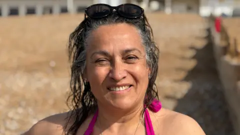 A dark-haired woman with wet hair in a pink swimming costume and sunglasses on her head 