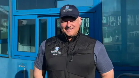 Cameron Weldon/BBC Saltash United Football Club sponsor Wayne Hambly stood in front of the old blue Tamar Bridge toll booth in its new home at Kimberley Stadium. He is wearing a blue hat and t-shirt with a black gilet. 