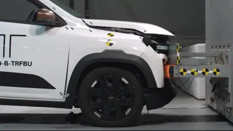 A white electric car crashing into a wall at a low speed in a crash test lab. The front of the car has bent in a bit and the bonnet has lifted up, but otherwise the damage looks minimal.