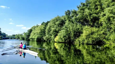 Elsie from the block Two rowers in a white boat on a river. The banks are lined with lush green trees and the sky overhead is blue. The water is still and the scene is brightly being reflected in the water.