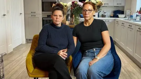 Renee Turner, left, has dark hair tied back and is wearing a navy jumper, black trousers, and is sitting on a yellow chair. Ruth O'Loughlin, right, has brown hair tied back, glasses, a black shirt and jeans and is sitting on a dark blue chair. They are both in a kitchen with units and cupboards behind them.