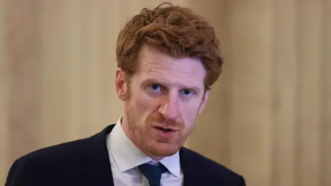 PA Media Matthew O'Toole  is wearing a suit with a white shirt and a tie. He has auburn hair. He's standing with white marble walls behind him.